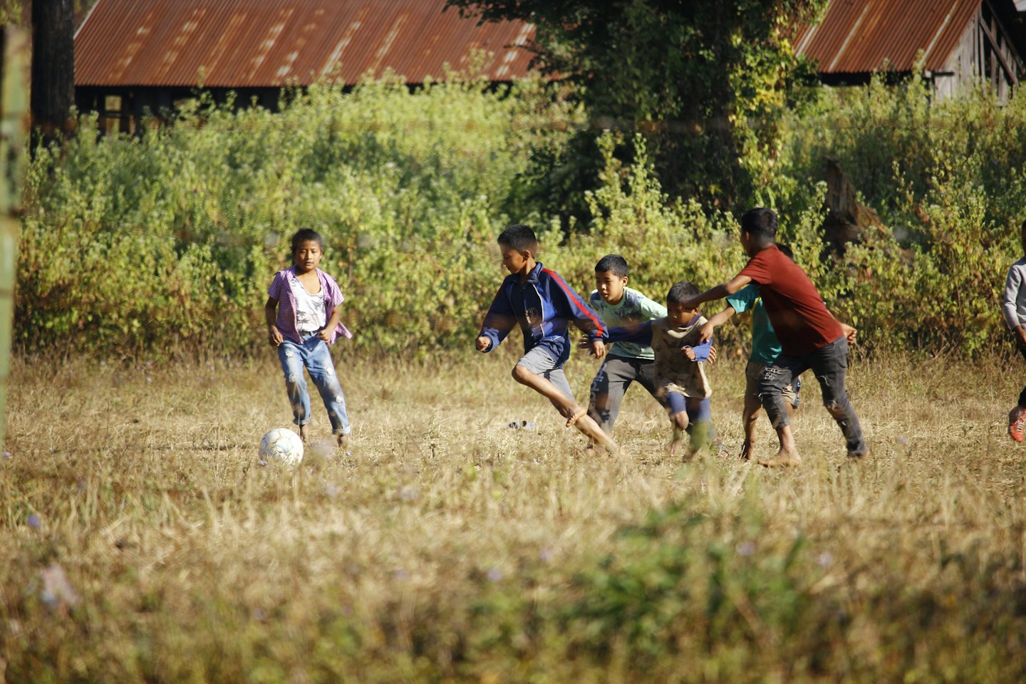 Bambini che giocano a calcio su un prato con edifici rustici.